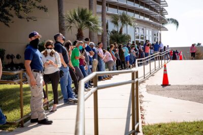 People wearing masks waiting in a line outside of a hospital.