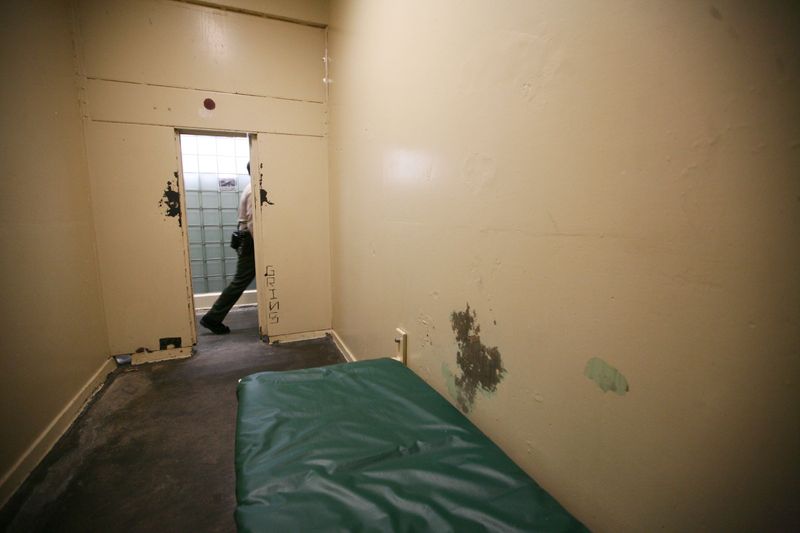 A cot with a plastic-looking green mattress in a narrow white room with patches of missing paint.