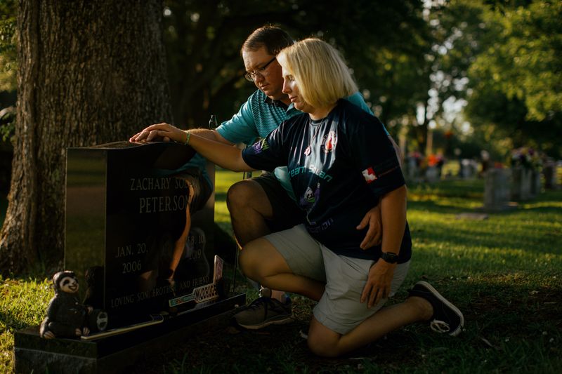 A white man and woman kneel before a grave, each with one hand on the top of the stone.