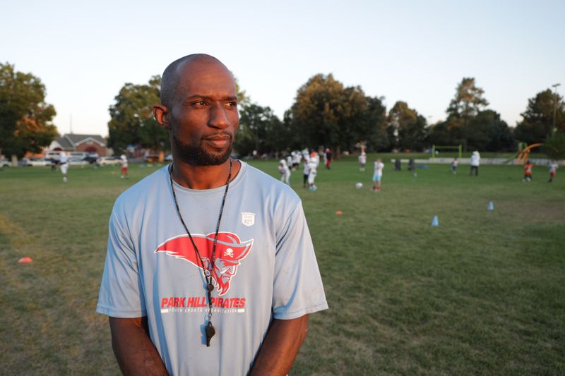 A bearded Black man wearing a T-shirt and a whistle stands in front of a field of children playing sports.