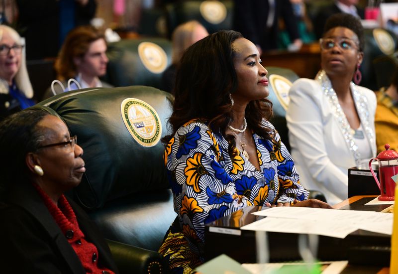 A Black woman in a flowered blue, orange and white dress sits in a large leather chair in the middle of a meeting.